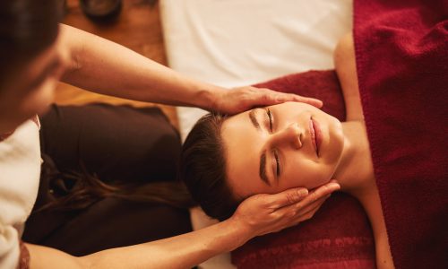 Calm Caucasian woman lying with closed eyes while massage practitioner putting palms on both sides of her face