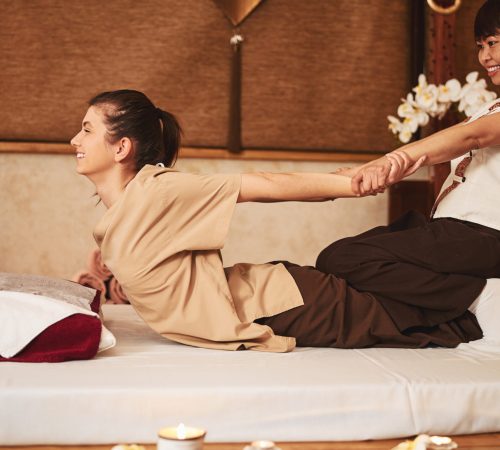 Lady lying on stomach on bed in spa and bending back while masseuse on top of her dragging her arms